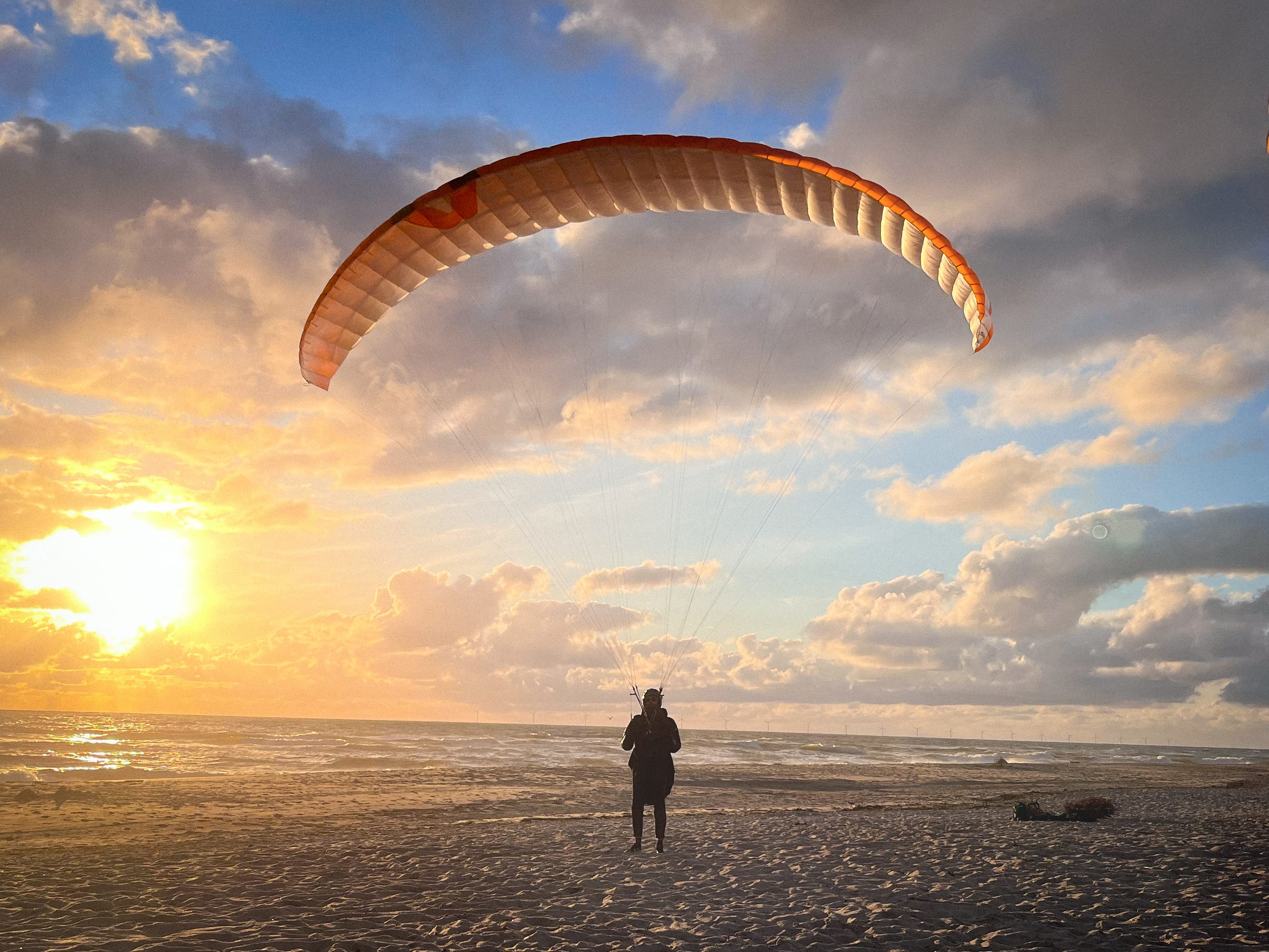 Ein Gleitschirmpilot steht in der Abenddämmerung am Strand, der Gleitschirm befindet sich über ihm in der Luft.